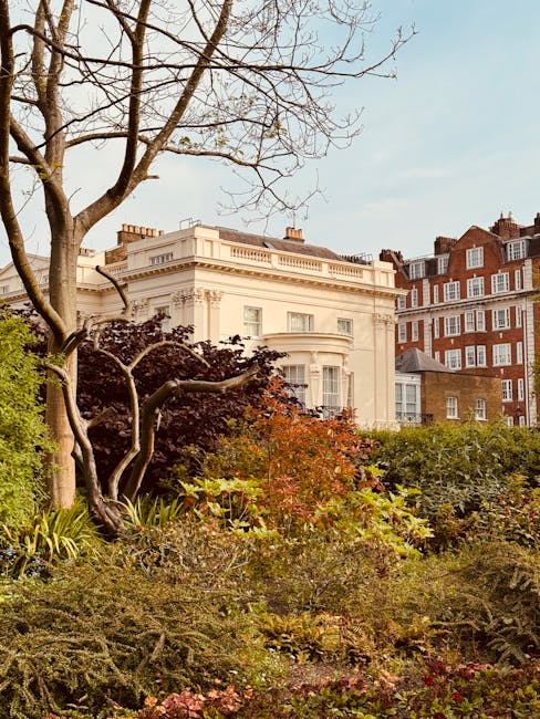 A lush, well-maintained garden in front of a stately white building with classical architectural details, including large windows and a decorative balustrade. The garden features a variety of plants,