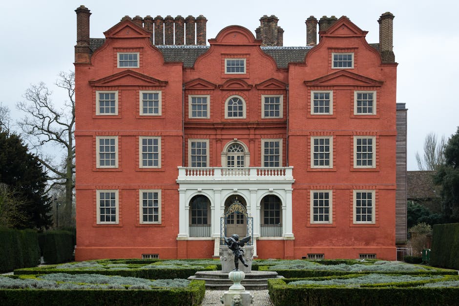 A detailed view of the well-maintained gardens in front of a historic brick building, featuring a lush green lawn with neatly trimmed grass, rounded garden beds with various plants, and a classical ma